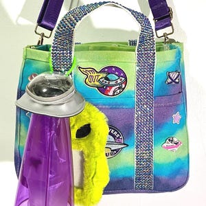 May include: A tie-dye tote bag in shades of green, blue, and purple with rhinestone-encrusted handles and a purple shoulder strap. The bag features space-themed patches, a fuzzy neon green alien charm, and a purple translucent UFO-shaped container.