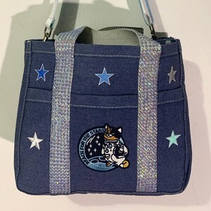 May include: Denim tote bag with a blue and white ombre strap and silver accents. The bag features embroidered stars and a patch with a cat in a space suit. The text on the patch reads "Reach for the Stars".
