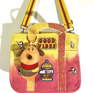 May include: A yellow and pink ombre canvas tote bag featuring a rhinestone-encrusted handle, a detachable plush dog burger charm, and embroidered patches including the text GOOD VIBES, Snoopy playing baseball, and Snoopy driving a car.