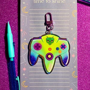May include: A holographic green, purple, and blue Nintendo 64 controller keychain with a green alien face on the front. The keychain has a silver ring and is attached to a gray notebook with the words "time to shine" written on it.