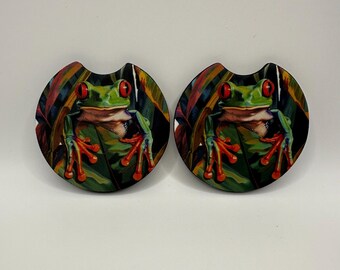 May include: Two car coasters featuring a vibrant illustration of a red-eyed tree frog. The frog is green with red eyes and orange feet, set against a backdrop of colorful leaves. These coasters are designed to fit in most car cup holders.