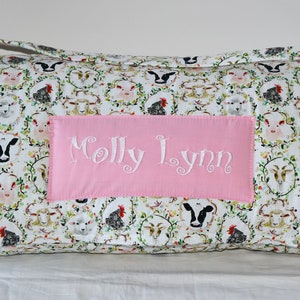 May include: A white and pink personalized nap mat with a farm animal print. The mat has a pink pocket with the name "Molly Lynn" embroidered in white.