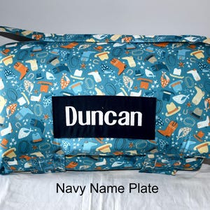 May include: A rolled fabric mat featuring a teal background with a repeating pattern of orange and white cowboy boots, hats, lassos, and stars. A rectangular navy blue patch is centred on the front with the name Duncan in white text.
