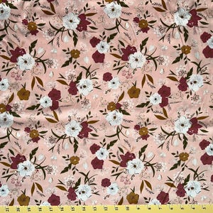 May include: Floral fabric with a repeating pattern of white, burgundy, and gold flowers on a light pink background. The fabric is decorated with green leaves and small white dots. The fabric is measured with a yellow ruler.