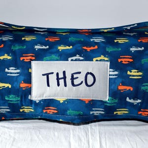 May include: A blue and white personalized nap mat with a white label that says "THEO" and a pattern of colorful airplanes on a blue background.
