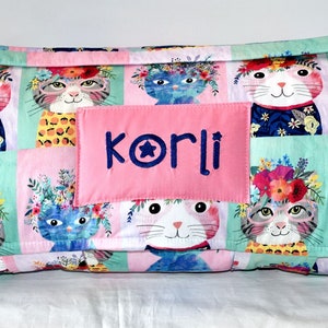 May include: A pink and green quilted nap mat with a patch that says "korli" in blue embroidery. The mat features a repeating pattern of cats wearing flower crowns.