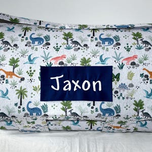 May include: A rectangular, patterned bag featuring a dinosaur theme. The bag is white with a repeating pattern of dinosaurs, trees, and plants in various colors. A navy blue rectangle with the name "Jaxon" in white script is centered on the bag.