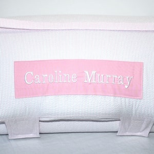 May include: A white and pink striped pillow with the name "Caroline Murray" embroidered in white on a pink rectangular patch. The pillow has a pink border and two pink fabric tabs.