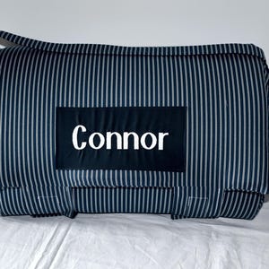 May include: A rectangular, navy blue and white striped bag with a handle. The word "Connor" is displayed in white on a black rectangular patch on the front of the bag. The bag is on a white surface.