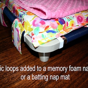 May include: A nap mat with a floral patterned cover and a pink blanket on top. The mat has elastic loops and is on a dark wooden surface. Text on the image reads: "Elastic loops added to a memory foam nap mat or a batting nap mat."