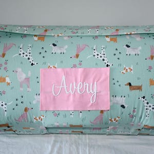 May include: A light green dog bed with a repeating pattern of various dog breeds and floral accents. A pink rectangular patch with the name "Avery" embroidered in white is centered on the bed.