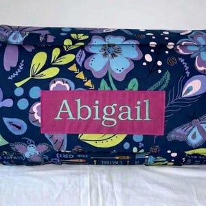 May include: A rectangular fabric mat with a dark blue background featuring a colorful floral and butterfly pattern in shades of light blue, purple, and lime green. A rectangular pink patch is centered on the front with the name Abigail embroidered.
