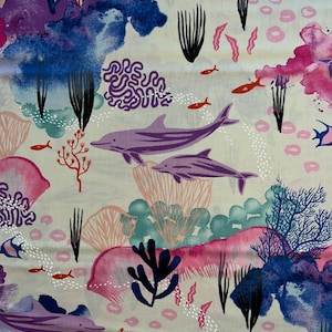 May include: Fabric with a marine life design. The pattern features purple dolphins, red fish, and coral in shades of pink, blue, and purple. The background is a light beige color. The design is reminiscent of an underwater scene.