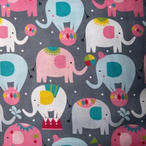 May include: A repeating pattern of circus elephants in pink, blue, and white on a gray background. The elephants are decorated with colorful accents and are balancing balls or standing on circus platforms. The design includes small white dots.