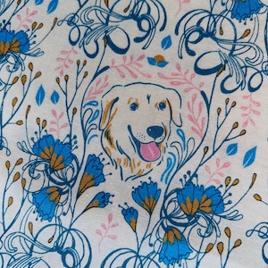 May include: White fabric with a repeating pattern of blue flowers, pink leaves, and a smiling golden retriever dog. The dog is surrounded by a pink floral wreath. The design has a whimsical, artistic style.