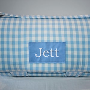 May include: A blue and white gingham patterned blanket or mat, with a blue rectangular patch embroidered with the name "Jett" in white. The blanket has a handle for carrying.
