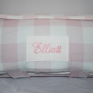 May include: Pink and white gingham fabric sleeping mat with a white patch sewn on the front with the name "Elliott" embroidered in pink.