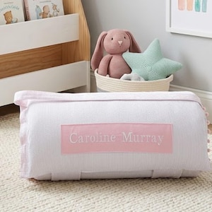 May include: A cylindrical bolster pillow featuring a white and light pink pinstripe fabric with a rectangular pink patch in the center. The patch is embroidered with the name Caroline Murray in white thread. The pillow has small pink pom-poms.