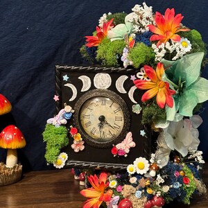 May include: A decorative black clock with a vintage-style face, surrounded by moss, flowers, and butterflies. The clock features a moon phase design and is adorned with miniature mushrooms and colourful floral accents.