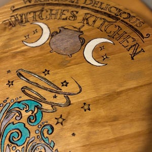Pode incluir: Tábua de servir de madeira com o texto "Wickedly Delicious Witches Kitchen". A tábua apresenta uma lua crescente, estrelas, um caldeirão e um design de redemoinho decorativo em turquesa, prata e branco. A tábua é de madeira com acabamento natural.