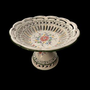 May include: White ceramic pedestal fruit bowl with a scalloped edge and intricate cut-out design. The bowl features a hand-painted floral pattern in various colors, with a green trim. The pedestal base is also decorated with floral motifs.