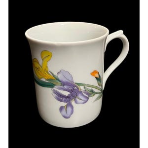 May include: White ceramic mug with a curved handle, decorated with a floral design of yellow, purple, and orange iris flowers. The flowers have green stems and leaves. The mug has a glossy finish against a black background.