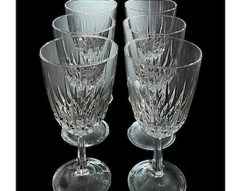 May include: A set of six clear crystal wine glasses. Each glass features a detailed, cut-glass pattern on the bowl and a slender stem. The glasses are arranged on a black background, showcasing their elegant design.