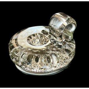 May include: A clear glass bottle stopper with a rounded top and a ring-shaped handle. The stopper has a textured, bubble-like design on its base and a smooth, reflective surface. The product is designed for use with bottles.