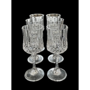May include: A set of six clear crystal wine glasses. Each glass has a detailed, cut-glass design on the bowl and a stemmed base. The glasses are arranged in two rows against a black background, highlighting their intricate patterns.