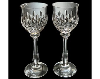 May include: Two clear crystal wine glasses with intricate leaf-like patterns on the bowls. The glasses have long, slender stems and round bases. The background is black, highlighting the clarity and detail of the glassware.