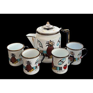 Op de afbeelding: Een witte emaille theeset met een theepot en vier mokken. De set heeft een ananasontwerp met bruine, blauwe en rode accenten. De mokken hebben gouden randen en zwarte handvatten. De theepot heeft een glazen knop.