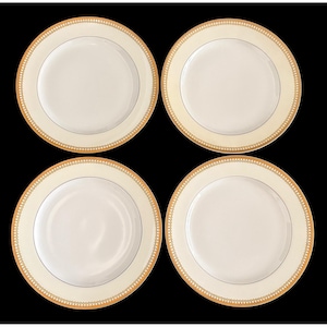 May include: Four white ceramic plates with a cream-colored border and a gold decorative trim. The plates have a simple, elegant design, suitable for formal dining. The plates are arranged on a black background.