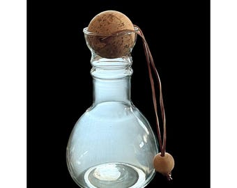May include: A clear glass bottle with a round base and a cork stopper. A brown leather cord and a wooden bead are attached to the stopper. The bottle is empty and set against a black background.