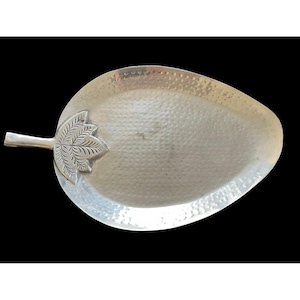 May include: Silver-colored, leaf-shaped serving tray with a textured surface. The tray features a detailed leaf design at the handle end. The tray is made of metal and is suitable for serving food or as a decorative piece.