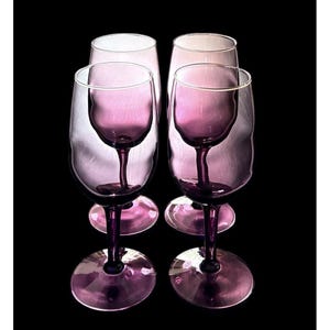 May include: A set of four wine glasses crafted from translucent purple glass. Each glass features a smooth, rounded bowl, a slender stem, and a circular base. The glasses are arranged in a two-by-two formation, showcasing the deep violet hue.