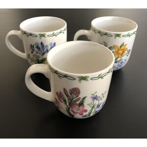 May include: Three white ceramic mugs with floral designs. Each mug features a different arrangement of colorful flowers, including blue, yellow, and pink blooms, and a green vine border. The mugs have curved handles.