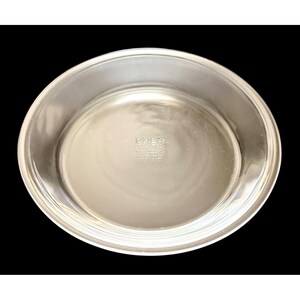 Może przedstawiać: Przezroczysta forma do ciasta Pyrex o okrągłym kształcie i lekko podniesionej krawędzi. Naczynie ma gładką, odblaskową powierzchnię i jest przeznaczone do pieczenia. Logo i tekst Pyrex są widoczne na spodzie naczynia.
