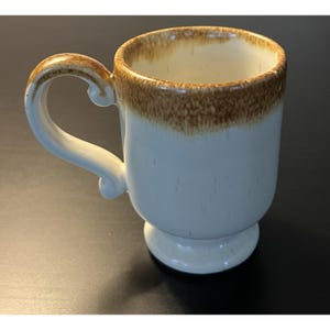 May include: A white ceramic coffee mug with a brown rim and handle. The mug has a pedestal base and a curved handle. The interior of the mug is white. The mug is on a black surface.