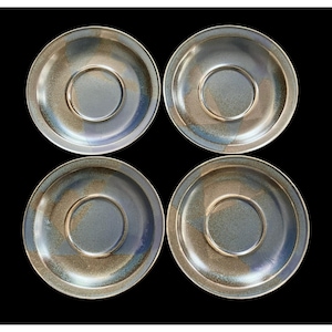 May include: Four ceramic saucers with a speckled, iridescent glaze in shades of gray, blue, and brown. Each saucer has a central recessed area and a raised outer rim. The saucers are arranged in a square formation against a black background.
