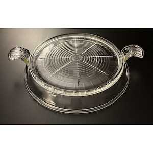 Puede incluir: Cazuela Fire King de vidrio transparente con tapa. El plato tiene dos asas y un diseño circular en la tapa. La tapa tiene una serie de círculos concéntricos y líneas radiales. El plato está sobre una superficie negra.