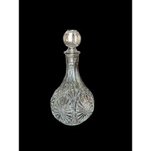 May include: A clear glass decanter with a round, patterned base and a long neck. The decanter has a decorative stopper. The glass is cut with a starburst pattern on the base and vertical lines on the neck. The background is black.