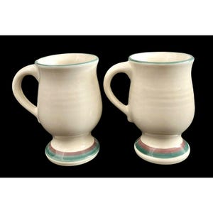 Può includere: Due tazze in ceramica bianco sporco con forma arrotondata e manico. I bordi sono rifiniti con un colore verde chiaro e la base presenta una fascia verde e marrone. Le tazze sono su sfondo nero.