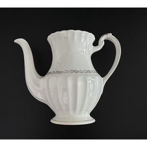 May include: A white ceramic teapot with a curved spout and handle. The teapot has a ribbed design and a decorative band around the middle. The rim and base are trimmed with a gold-colored accent. The background is black.