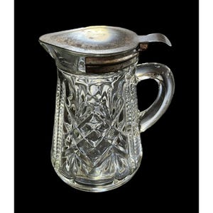 May include: A clear glass creamer with a diamond pattern and a silver-colored lid. The creamer has a handle and a spout for easy pouring. The lid is hinged and can be opened and closed. The creamer is likely vintage and suitable for serving cream or other liquids.