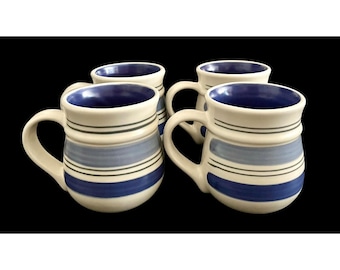 May include: Four ceramic mugs with a cream-colored base, blue and gray horizontal stripes, and a dark blue interior. Each mug has a curved handle and a rounded shape. The mugs are arranged against a black background.