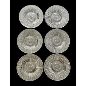 May include: Six clear glass saucers with a detailed wheat stalk design radiating from a central starburst pattern. The saucers are arranged on a black background, showcasing their intricate embossed details. A classic tableware set.