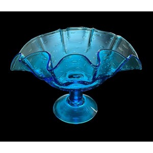 May include: A vibrant blue glass bowl with a ruffled edge and a pedestal base. The bowl has a textured surface with small air bubbles, adding a unique visual element. The bowl is a decorative piece.