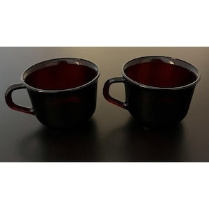 Puede incluir: Dos tazas de café negras con interior rojo y borde negro. Cada taza tiene un asa curva y un borde ligeramente inclinado. Las tazas están sobre un fondo oscuro.