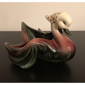 Peut inclure: Un pot de fleurs en céramique en forme de canard, avec une tête blanche et un corps rouge et vert. La tête du canard est ornée d'un foulard blanc à pois rouges. Le pot a un design incurvé, avec une large ouverture pour les plantes ou autres objets décoratifs.