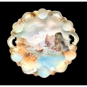 Puede incluir: Un plato decorativo de porcelana con una escena de paisaje marino pintada a mano. El plato presenta un borde festoneado con ribete dorado y varias hendiduras circulares. La obra de arte representa un velero en el agua, acantilados y montañas bajo un cielo nublado.
